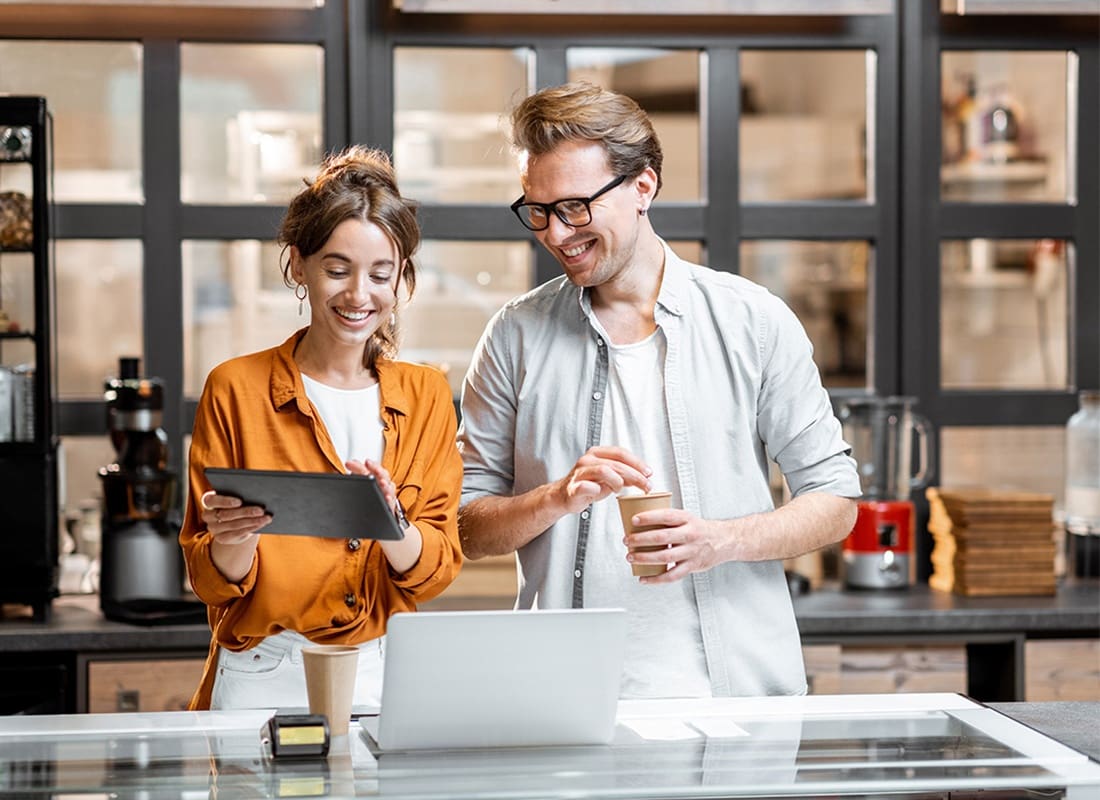Business Insurance - Cheerful Young Female and Male Small Business Owners Standing Behind the Front Counter of Their Cafe Looking at a Digital Tablet