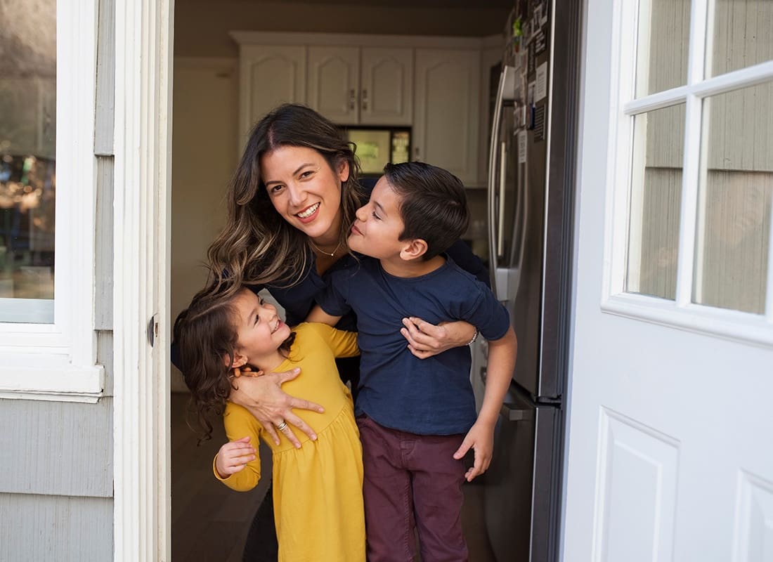Insurance Solutions - Portrait of a Cheerful Young Mom Hugging her Son and Daughter While Standing in the Front Doorway of their Home